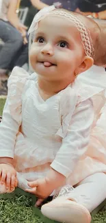 Adorable baby sitting on grass in a cute white dress and smiling.