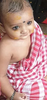 Smiling baby in red and white traditional attire on a colorful mat.