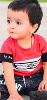 Adorable baby in red shirt sitting on ground with green background.