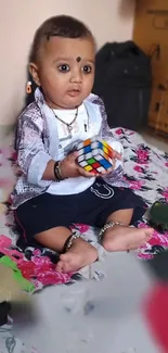 Adorable baby holding Rubik's cube on colorful floral mat.