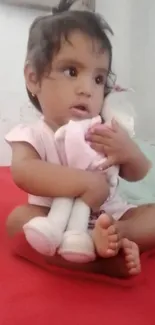 Adorable baby holding a stuffed toy while sitting on a red blanket.