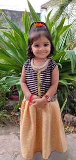 Smiling child in garden with green leaves and traditional attire.