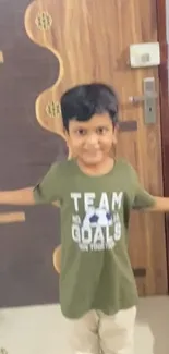 Smiling child in green t-shirt stands happily by a wooden door.