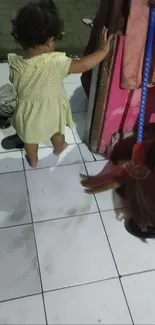 A toddler in a yellow dress in a colorful room setting with a broom nearby.