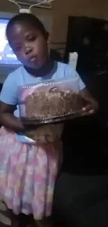 Adorable child holding a cake in a dimly lit living room.