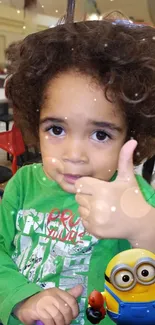 Adorable child with green shirt and a Minion character giving thumbs up.