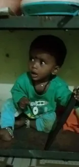 Adorable young child in green shirt sitting on a shelf, looking surprised.