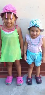 Two smiling kids in colorful outfits and hats against a brick wall.