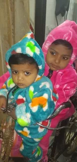 Two young children in colorful fleece outfits on a bike indoors.