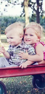 Two smiling kids on a red wagon outdoors.