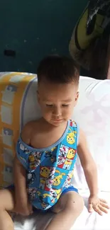 Adorable toddler in cartoon onesie sits on a colorful bed.