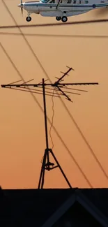 Airplane flying over silhouette of rooftop antenna at sunset.