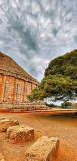 Ancient ruins beside a lush tree under a cloudy sky.