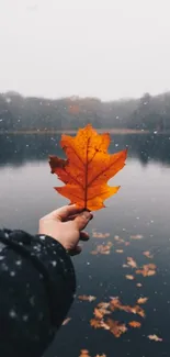 Hand holds orange leaf by a snowy lake, perfect for mobile wallpaper.