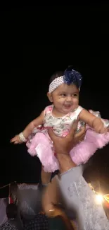 A joyful baby lifted high wearing a floral dress and smiling at an event.