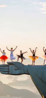 Ballet dancers gracefully pose on a statue's hand against a sunset sky.