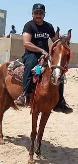 Man riding a brown horse on a sandy beach under a clear blue sky.