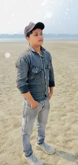 Young man in denim stands on a sandy beach.