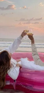 Two people relaxing on a beach in a pink inflatable pool, toasting at sunset.