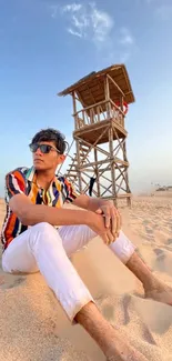 Man in colorful shirt relaxing on sandy beach with lifeguard tower.