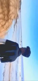 Person walking along a beach under a bright blue sky.