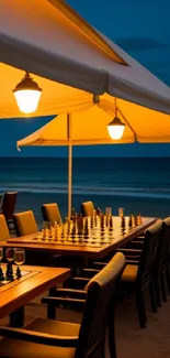 Chess tables under glowing umbrellas by the beach at dusk.