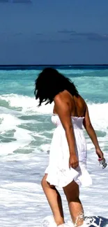 Woman in white dress walking on a sandy beach with blue ocean waves.