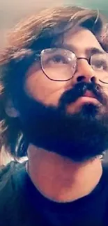 A bearded man with glasses, wearing a black shirt, looks upwards against a blurred background.