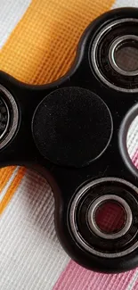 Black fidget spinner on orange and pink striped fabric background.