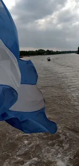 Blue and white flag waving over a river under a cloudy sky.