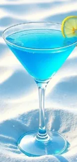 Blue cocktail in a martini glass on white sand beach.