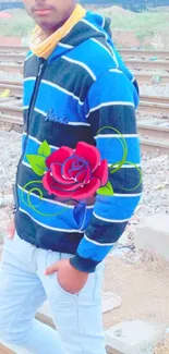 Person in blue hoodie with red rose by railway tracks.