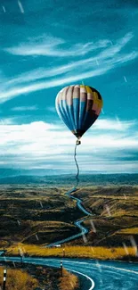 Hot air balloon floats over a winding road and landscape under a blue sky.