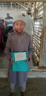 Young boy in rustic setting holding a blue book, surrounded by rural community.