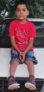 Young boy in red shirt sitting on a white car hood with trees in the background.