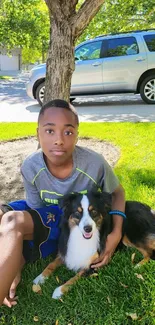 Boy sitting with dog on green grass under a tree, on a sunny day.