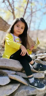 Young girl sitting on stones in sunlight wearing a yellow shirt.