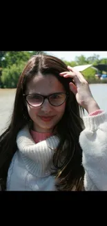 Woman with glasses smiling by a river in a cozy sweater.