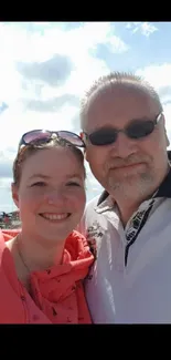 A couple smiling under a bright sky with scattered clouds.