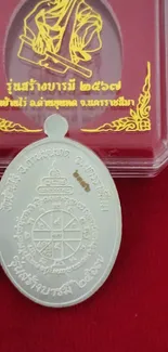 Silver Buddhist amulet on red cloth backdrop with decorative packaging.