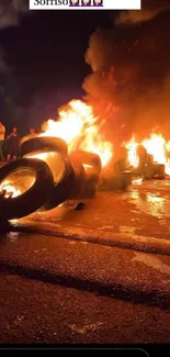 Burning tires on a dark road at night, with vivid flames and onlookers present.