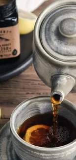 Teapot pouring tea and honey with lemon slices on a wooden table.