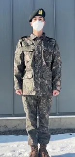 Person in camouflage uniform stands on snow with a gray industrial background.