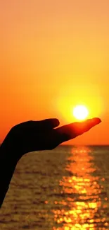 Silhouette of a hand holding the sun against an orange sunset over the ocean.