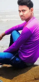 Man in purple shirt sitting on beach rocks.