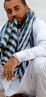 Man in white attire with blue scarf sitting by the beach.