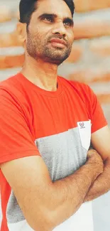 Man in red shirt against brick wall, arms crossed.