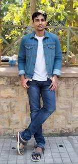 Man in denim standing outdoors with greenery in background.