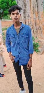 Young man in denim stands casually outdoors by a large tree.