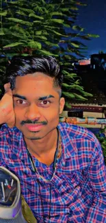 Young man sitting in front of greenery, wearing a checked shirt.